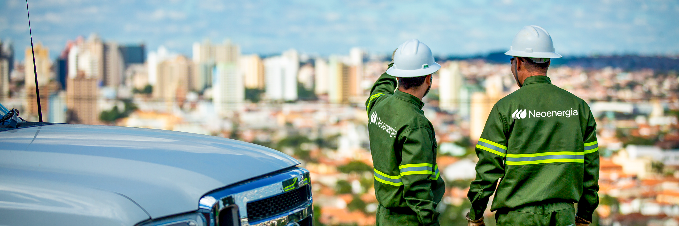 Dois eletricistas da Neoenergia próximos a viatura da empresa em local aberto observando o ambiente.